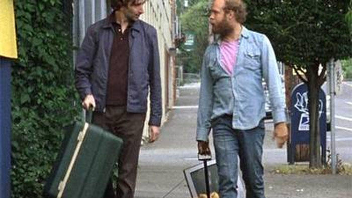 Two men are walking along the road talking. Both are holding suitcases or luggage type baggage.