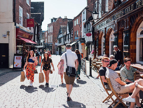 Several people are shopping and sitting outside cafes on a cobbled street in the sunshine
