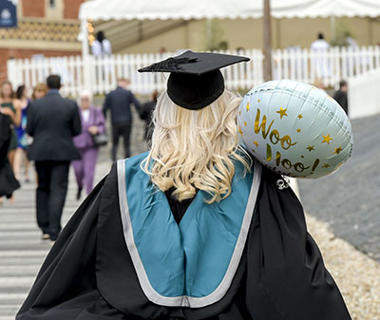 A student dressed in a graduation robe is facing away from the camera. She is holding a balloon that says "Woo Hoo" on it in gold lettering
