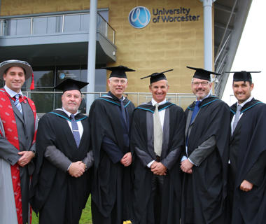 Group photo of mature men wearing graduation robes outside the university arena