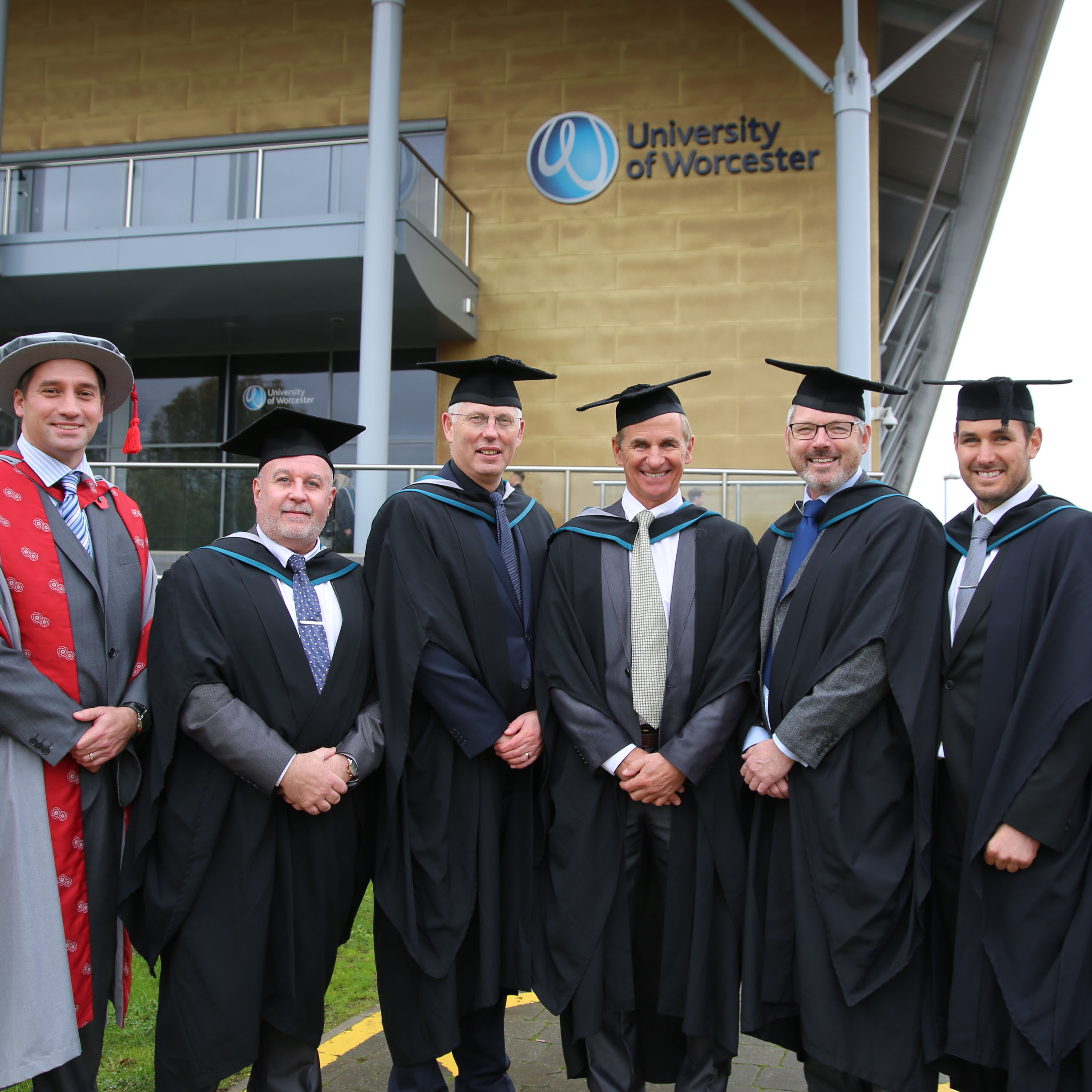 Group photo of mature men wearing graduation robes outside the university arena