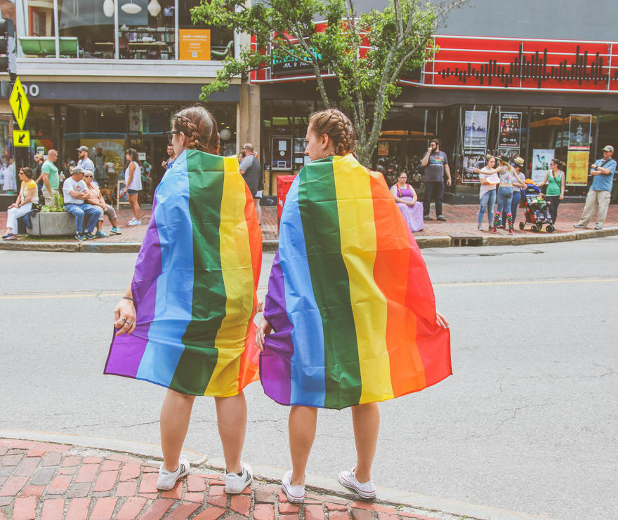 Two women are wearing the LGBTQ+ Pride Flag as capes