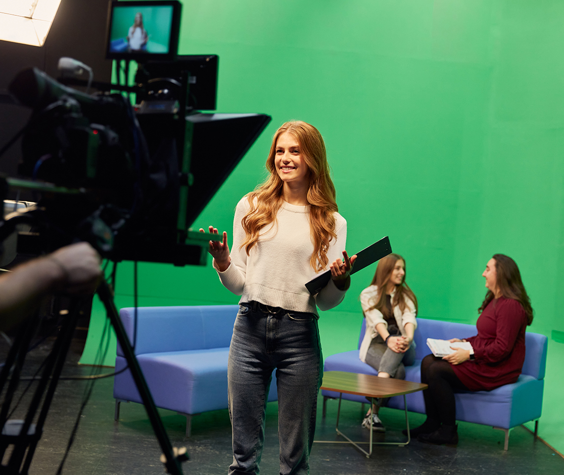 A girl stood in front of a film camera with two people in the background