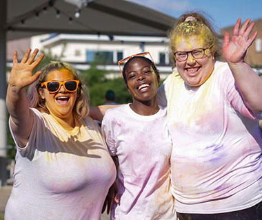 three smiling students are waving at the camera. They are covered in bright colours from the colour run.